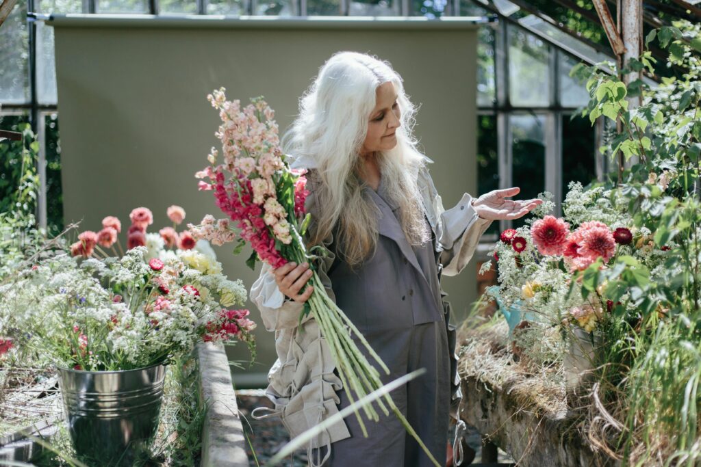 Senior woman arranging vibrant flowers in a greenhouse filled with various blooms.